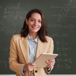 Waist up portrait of young female teacher looking at camera and smiling while standing by blackboard in school classroom, copy space